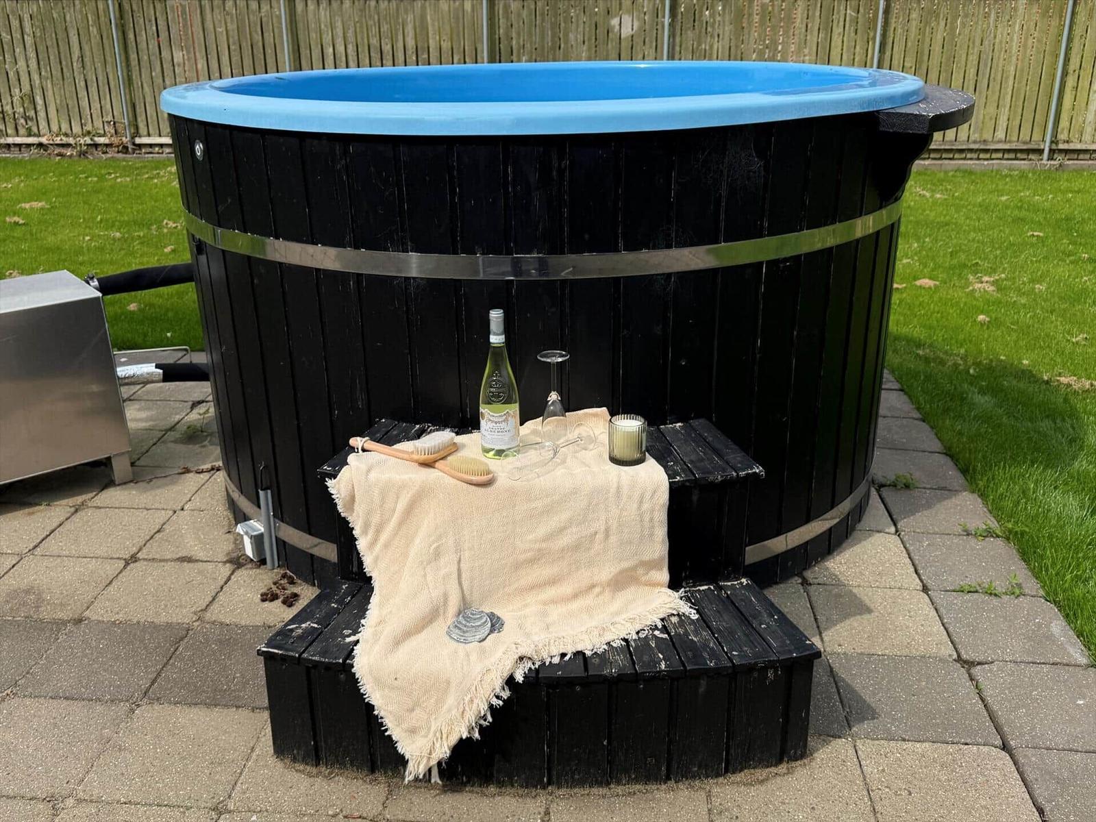 Ein schwarzer Holzwhirlpool mit blauem Innenrand. Davor steht eine Bank mit Tischdecke, Wein, Glas und Kerze.