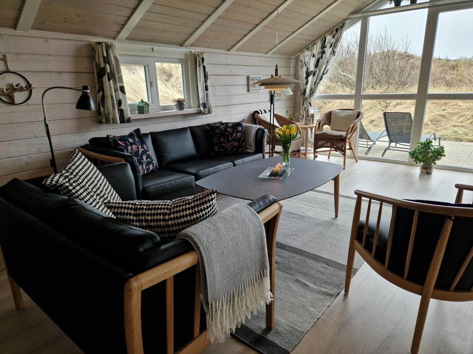 Wohnbereich mit Sofa, Tisch und Sessel. Große Fenster zeigen Landschaft.