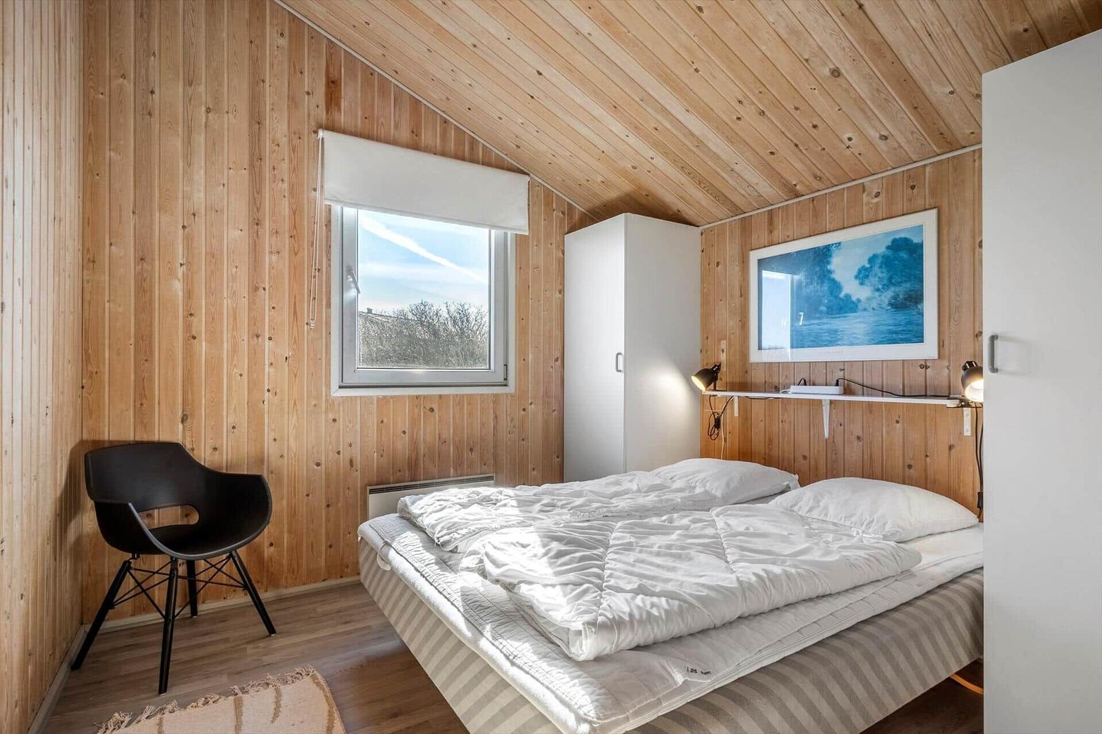 Schlafzimmer mit Holzwänden, Bett, Fenster und schwarzer Sitzgelegenheit.