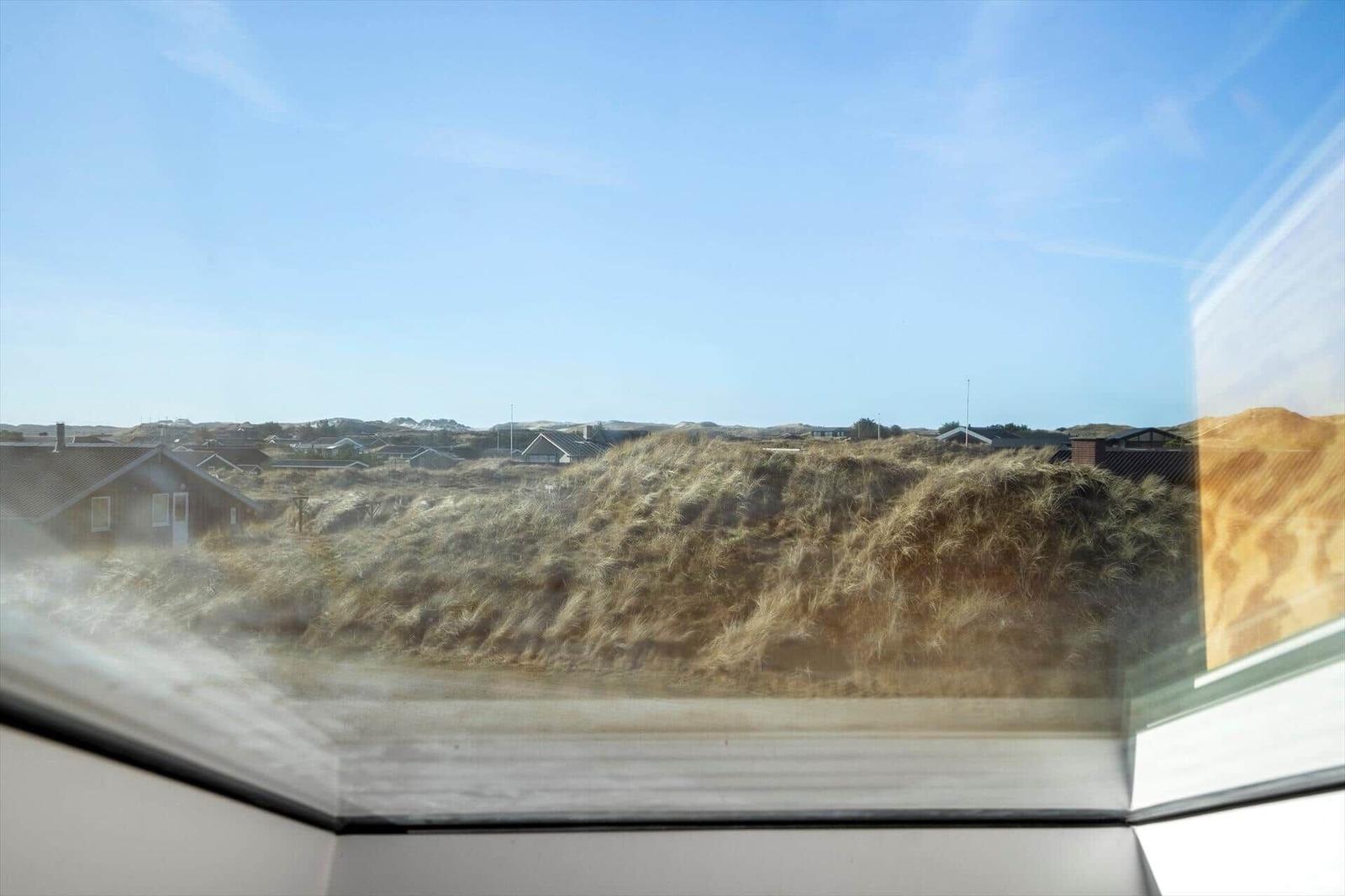 Blick aus Fenster auf Dünen mit Gras und Häusern im Hintergrund unter blauem Himmel.