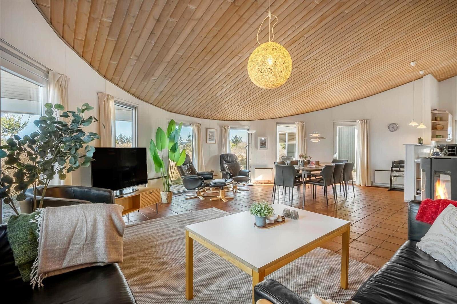 Großer Wohnbereich mit Holzdecke, Sitzgelegenheiten und Esstisch. Kamin und große Fenster.