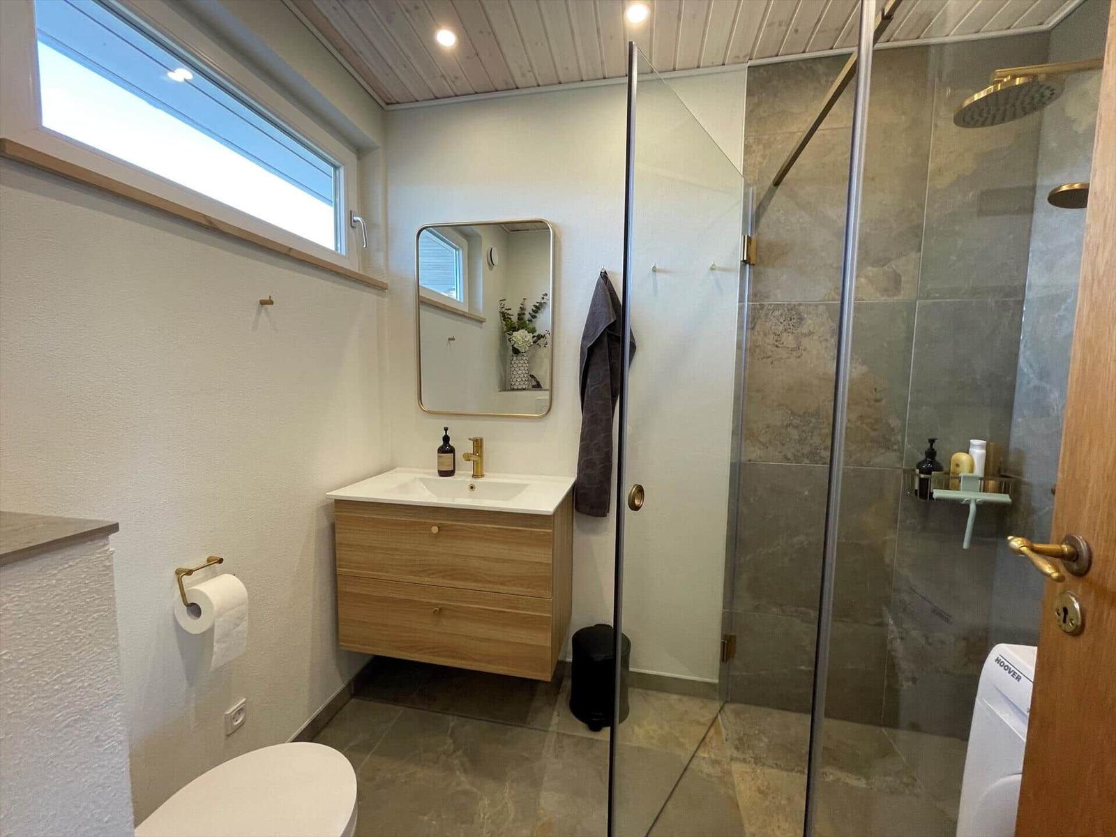 Badezimmer mit Dusche, Waschbecken und Toilette. Holzschrank, Spiegel und Fenster.
