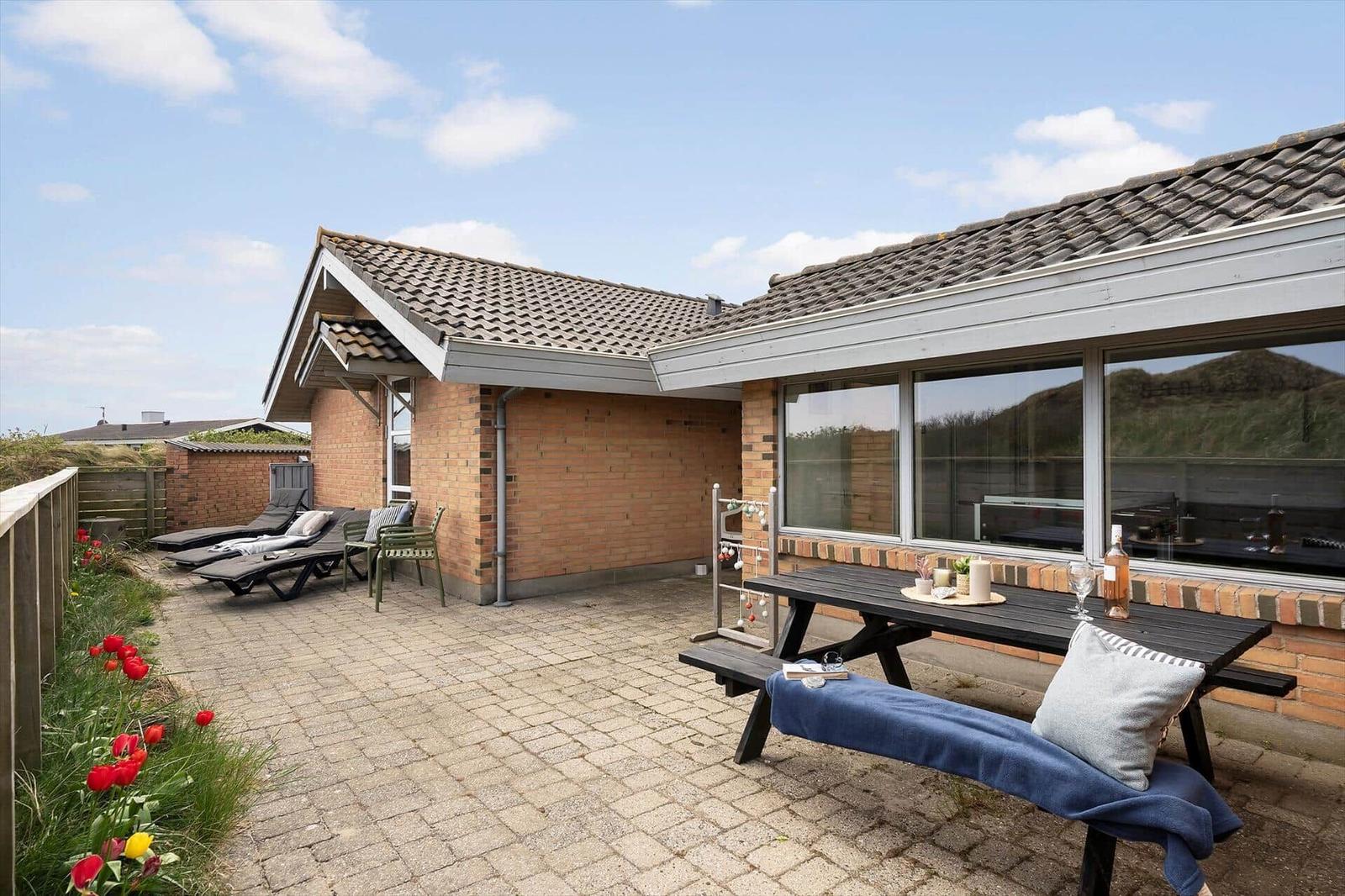 Terrasse mit Liegestühlen, Tisch und Blumenbeet. Haus mit Backsteinwand und Glasfront.