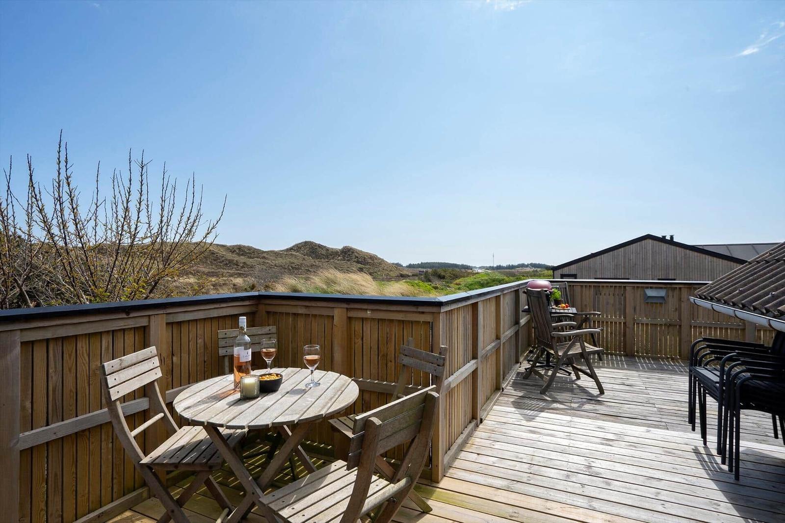 Holzterrasse mit Tisch, Stühlen und Grill. Blick auf Hügel und Himmel.