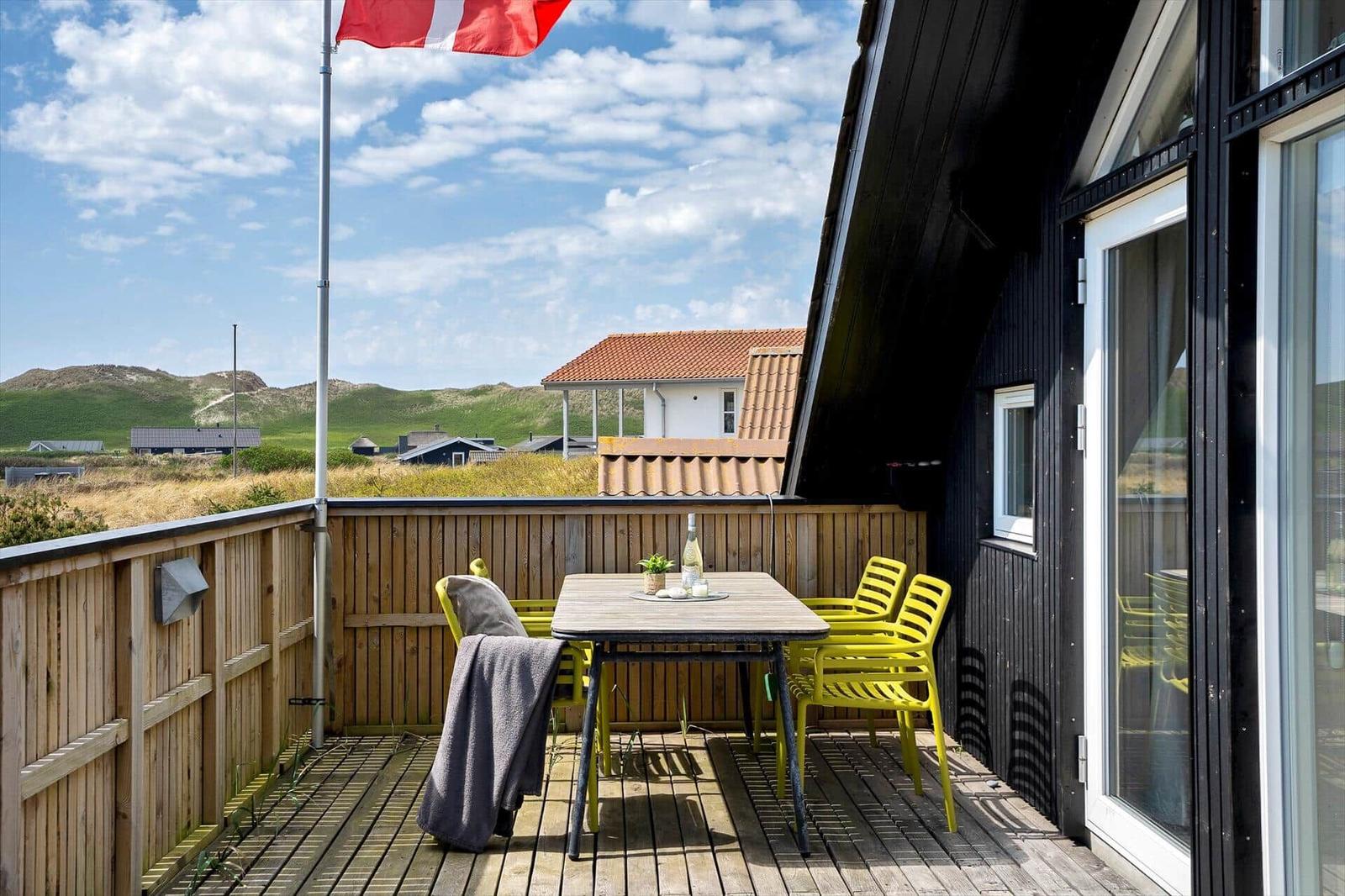 Terrasse mit Tisch, gelben Stühlen und dänischer Flagge. Blick auf grüne Hügel und ein Haus.