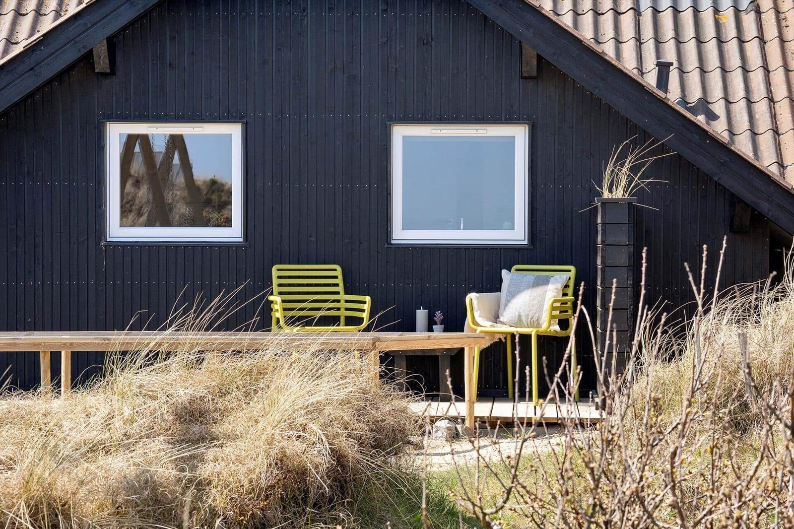 Schwarzes Haus mit zwei Fenstern und gelben Stühlen auf einer Holzterrasse.