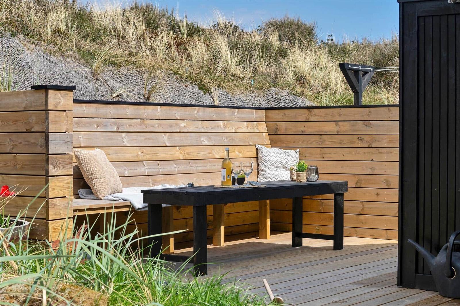 Holzterrasse mit Sitzbank, Tisch und Getränken vor Dünenhintergrund.
