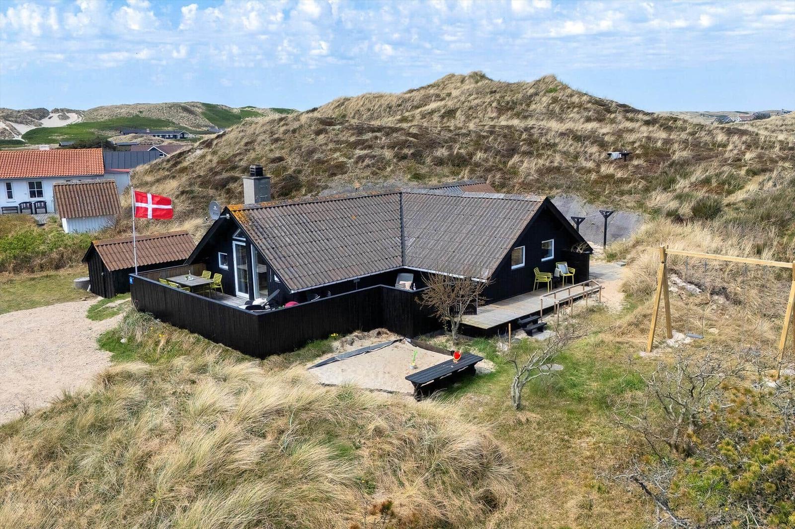 Schwarzes Ferienhaus mit Terrasse und Dachterrasse in Dünenlandschaft. Daneben steht eine Bank und ein Spielgerüst.