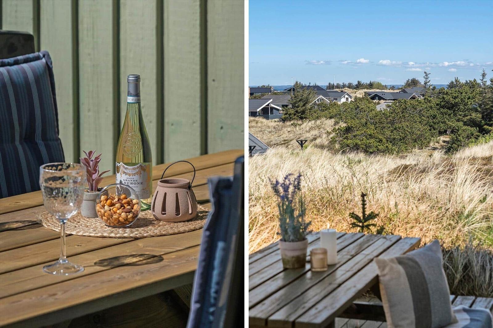 Holztisch mit Wein, Nüssen und Kerze auf Terrasse. Blick auf Hügel mit Häusern und Strand.