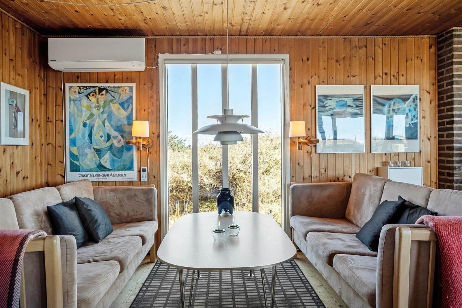 Wohnzimmer mit Holzwänden, Sofa, Tisch und Fenster. Wandbilder und Deckenventilator sichtbar.