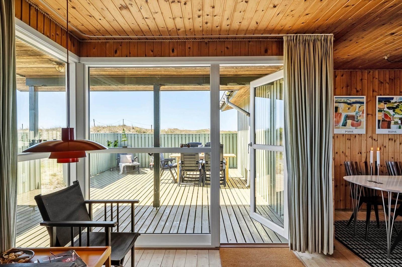 Wohnraum mit Blick auf Terrasse und Garten. Holzdecke, Fenster und Stühle sichtbar.