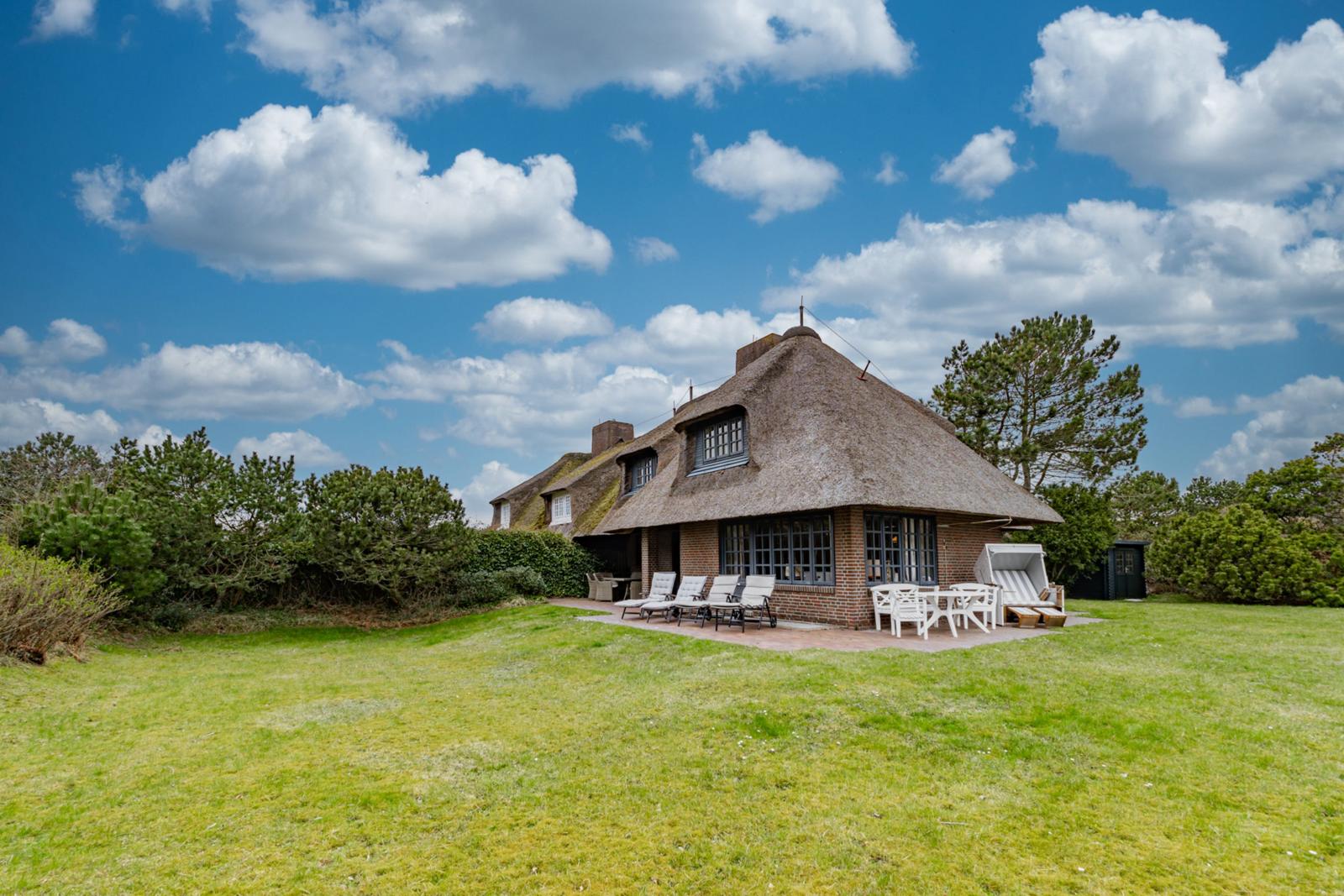 Haus mit Strohdach, Terrasse mit Liegen und Tisch, umgeben von Gras und Bäumen unter blauem Himmel.