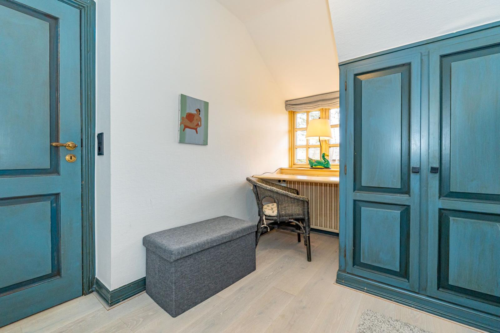 Ein Raum mit blauer Tür, grauem Sitzkasten und Schreibtisch unter dem Fenster.