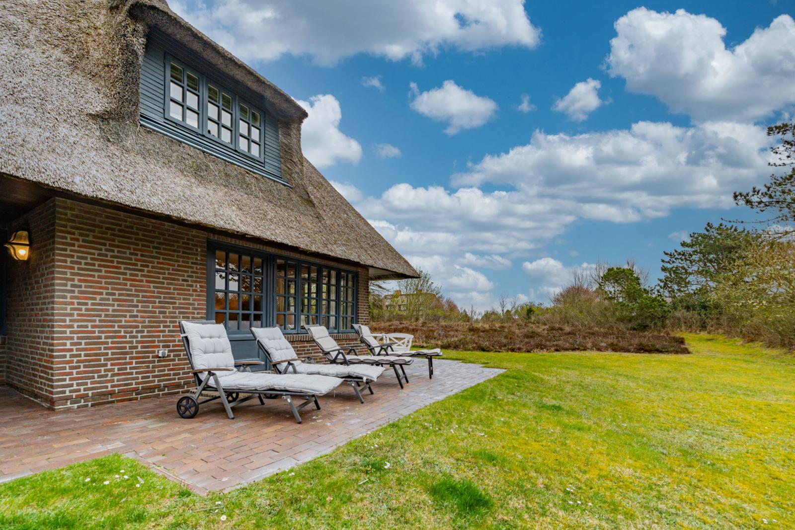Haus mit Strohdach, Terrasse mit Liegestühlen und grünem Rasen unter blauem Himmel.