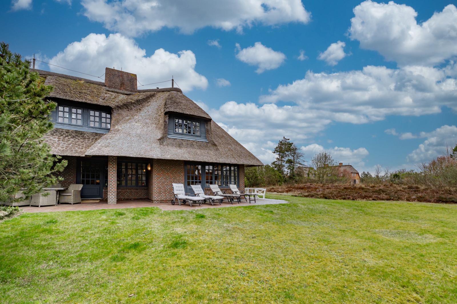Haus mit Strohdach, Terrasse und Liegestühlen auf grünem Rasen unter blauem Himmel.