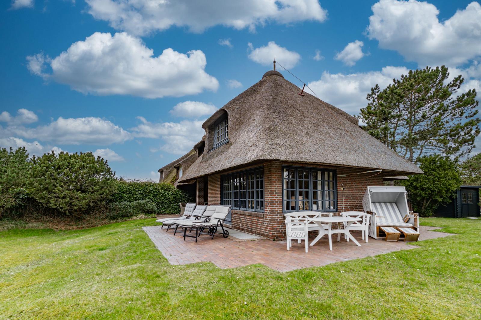 Dorfhaus mit Strohdach, Terrasse mit Tisch, Stühlen und Liegestühlen auf grünem Rasen.