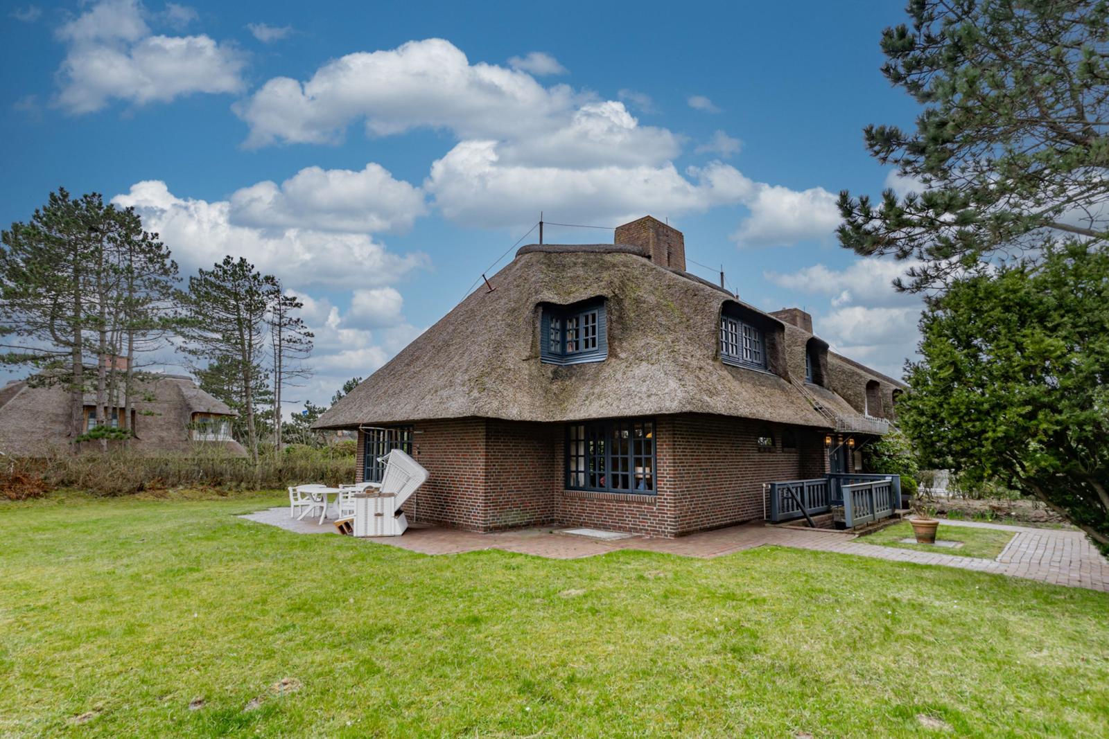Haus mit Strohdach, Garten und Terrasse unter blauem Himmel.