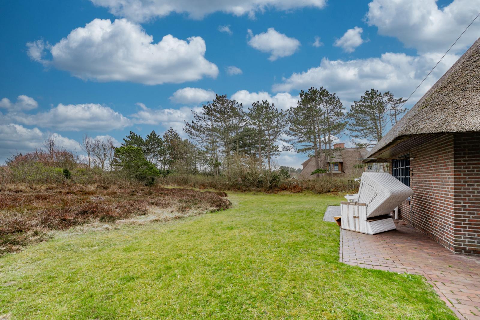 Haus mit Dach aus Stroh, Terrasse mit Liegestuhl und grünem Rasen. Hintergrund mit Bäumen und blauem Himmel.