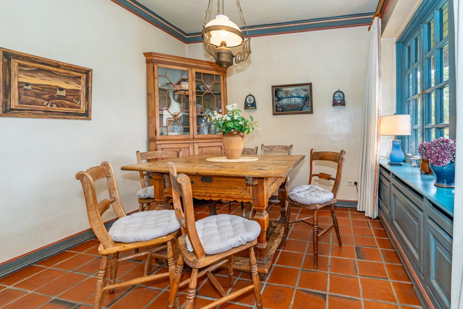 Esszimmer mit Holztisch, Stühlen und Terrakottboden. Fenster und Schrank sind sichtbar.