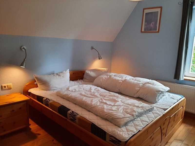 Schlafzimmer mit Holzbett, Nachttisch und Fenster. Decke ist schräg. Wandfarbe ist hellblau.