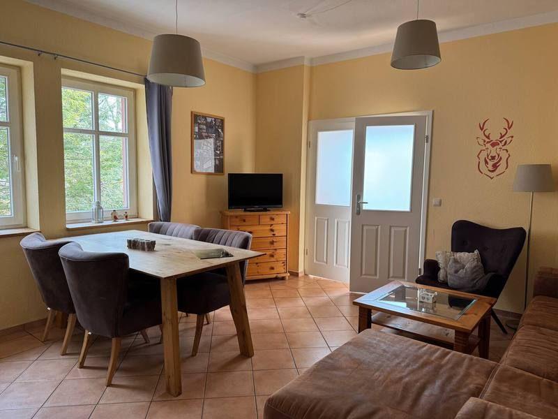 Essbereich mit Tisch und Stühlen, Wohnbereich mit Sofa und Couchtisch, Fernseher und Tür.