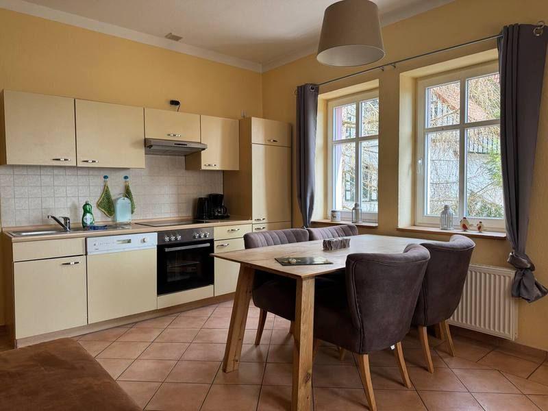 Küche mit Essbereich, Holztisch und Stühlen, Fenster mit Vorhängen und Küchenspüle.