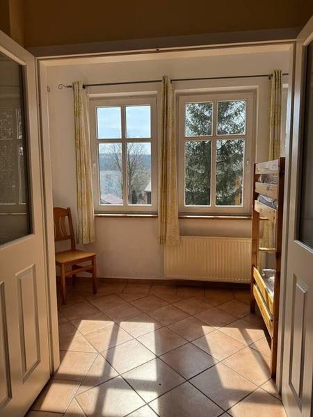 Raum mit zwei Fenstern, Holzstuhl und Kachelboden. Blick auf Wald und Landschaft.