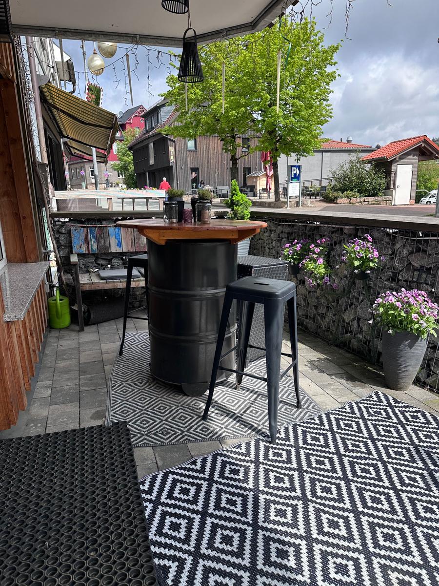 Terrasse mit Tisch, Stühlen und Blumen. Hintergrund zeigt Straßenszene mit Gebäuden.
