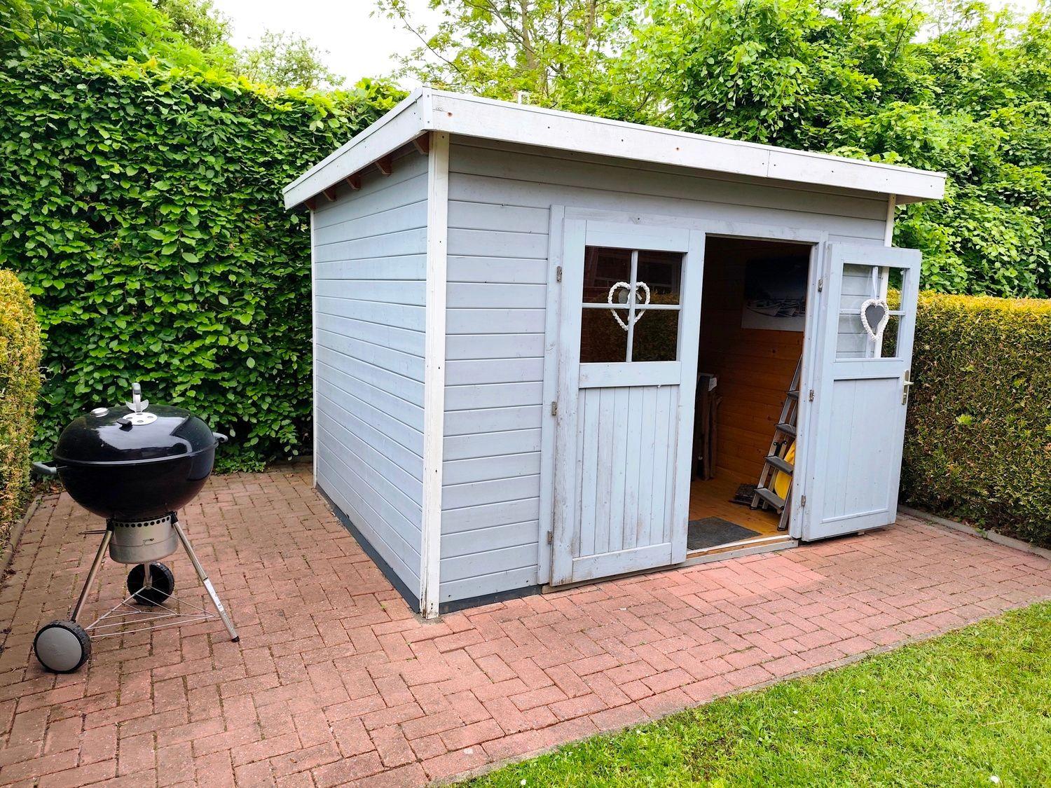 Gartenhaus mit Grill auf gepflastertem Platz zwischen Hecken.
