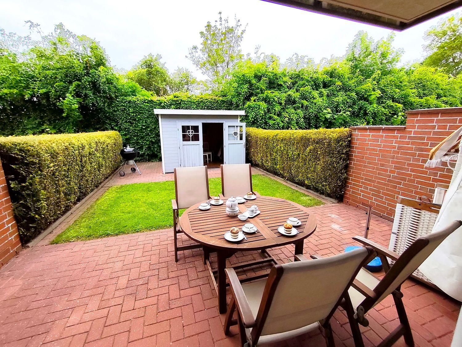 Terrasse mit Tisch, Stühlen und Grill. Hinterhof mit Gartenhaus und Hecke.