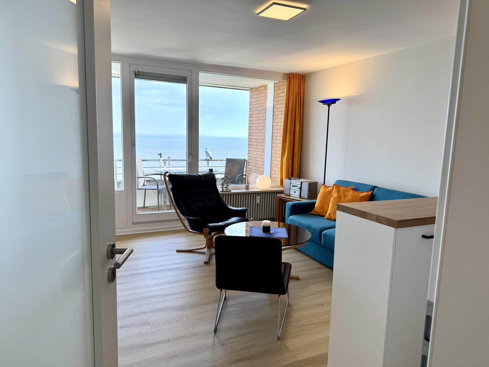Wohnzimmer mit Blick aufs Meer, blaues Sofa, schwarze Sessel und Holzboden.