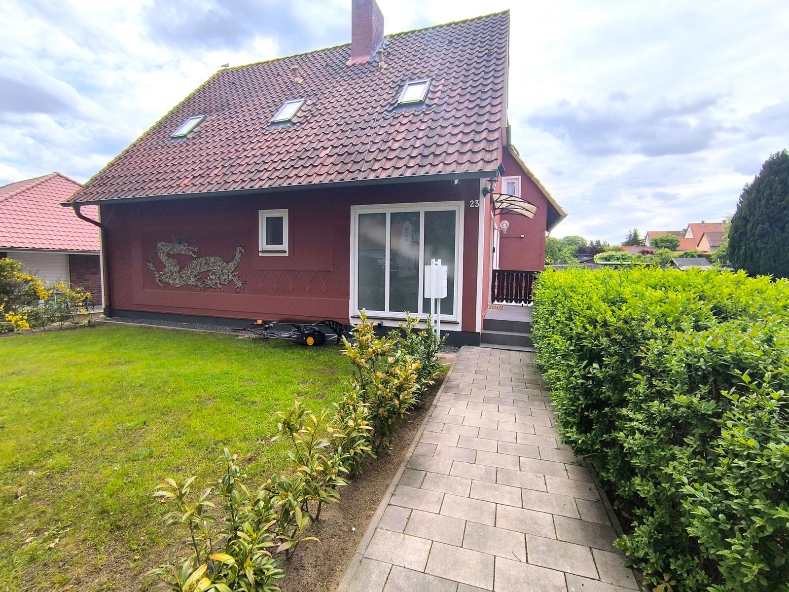 Rotes Haus mit Garten, Terrasse und Dachfenstern. Pfad führt zur Eingangstür.
