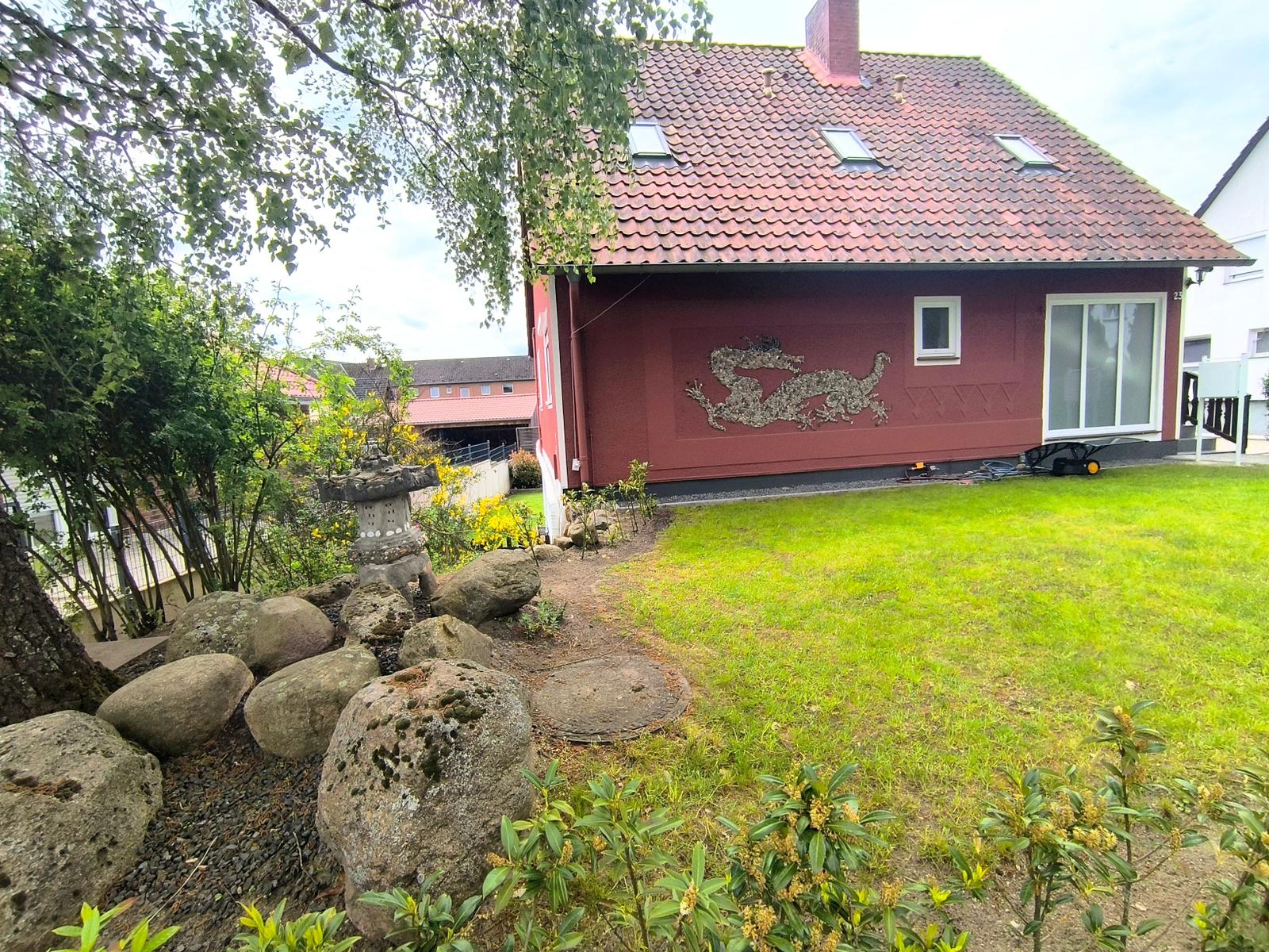 Rotes Haus mit Drachen-Mosaik, grüner Rasen und Steinbereich im Vorgarten.