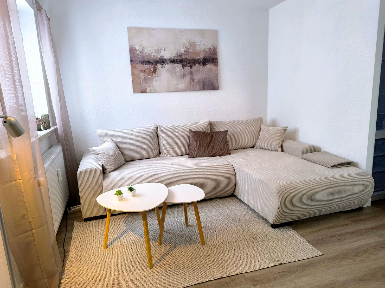 Wohnzimmer mit Ecksofa, zwei Tischen und Wandbild.
