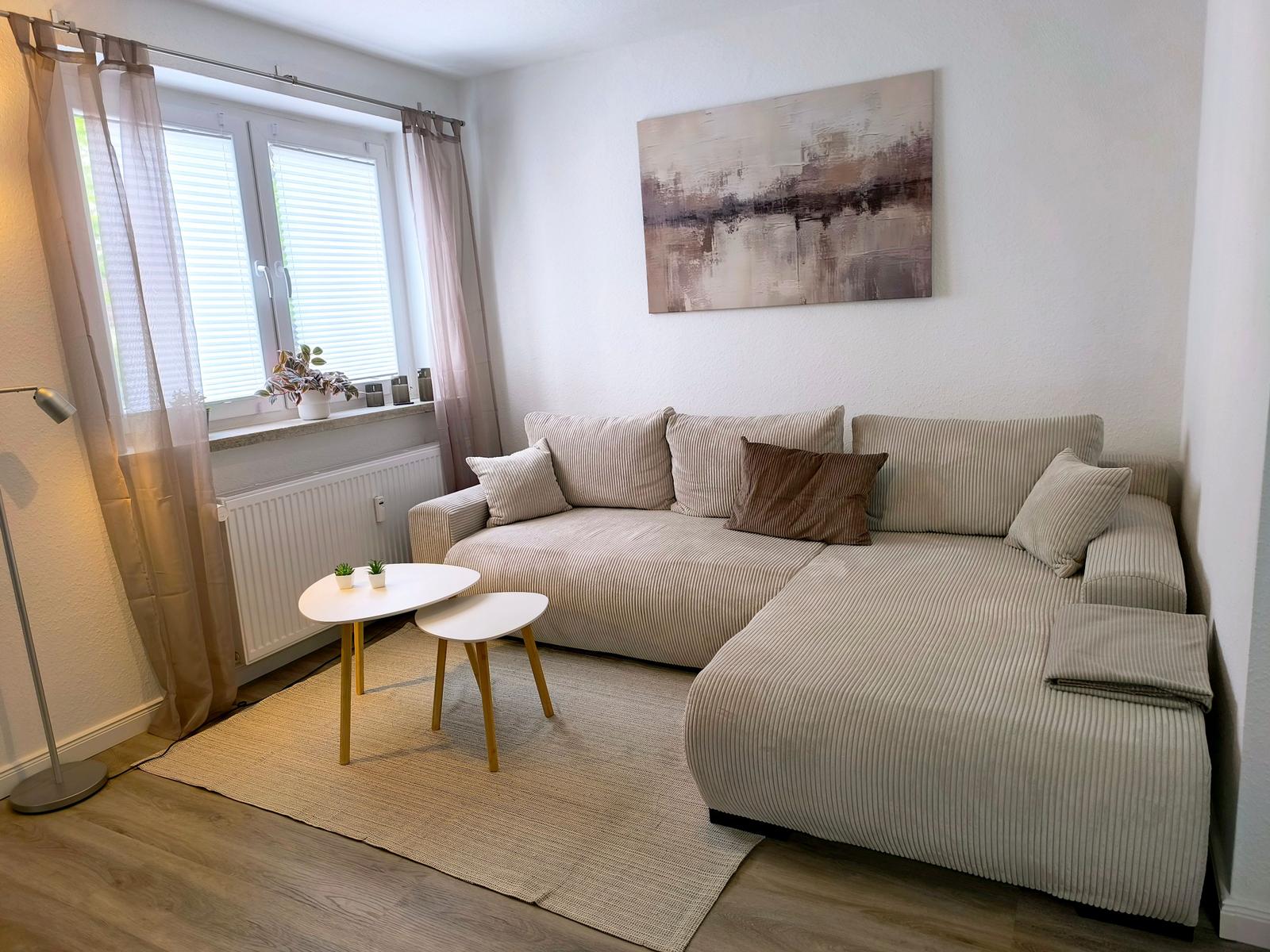 Wohnzimmer mit Ecksofa, zwei Tischen und Wandbild. Fenster mit Jalousien und Vorhängen.
