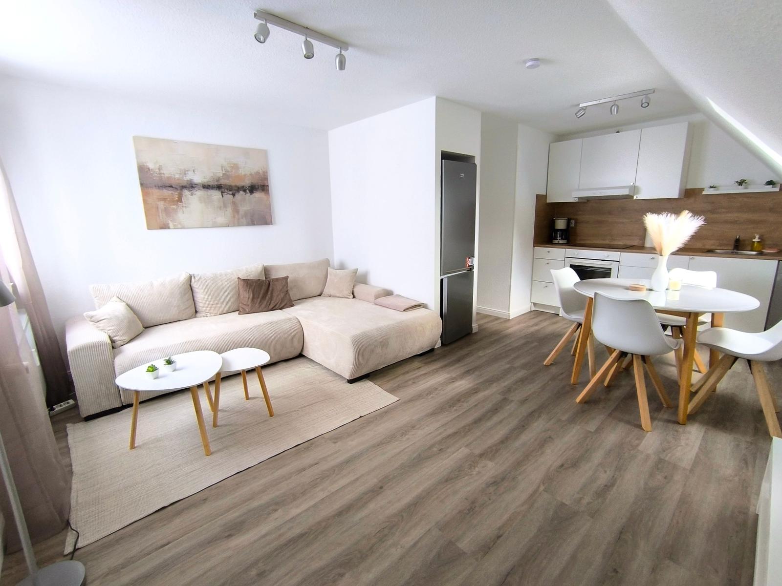 Wohn- und Essbereich mit Sofa, Esstisch und Küchenzeile. Holzdielenboden und weiße Wände.