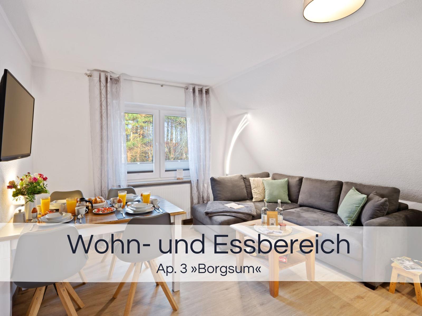 Wohn- und Essbereich mit Sofa, Tisch und TV. Fenster mit Blick auf Wald.