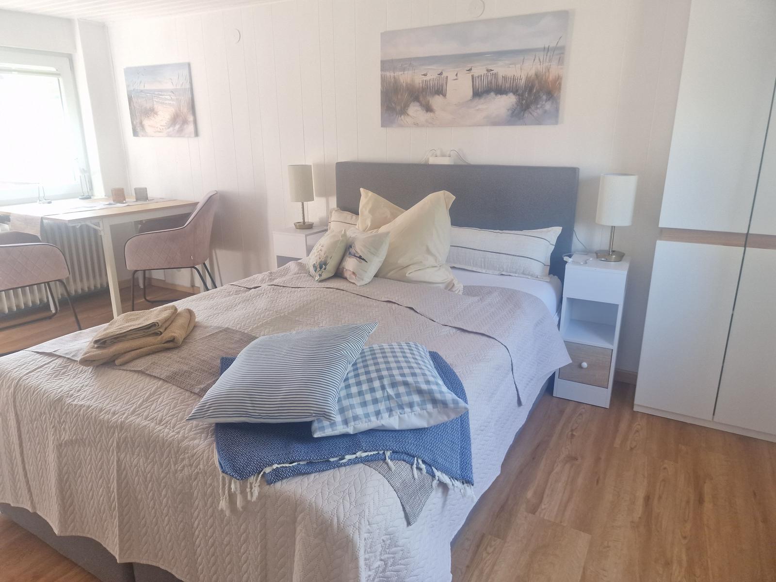 Schlafzimmer mit Bett, Nachttischen und Schreibtisch. Wandbilder zeigen Strandlandschaften.