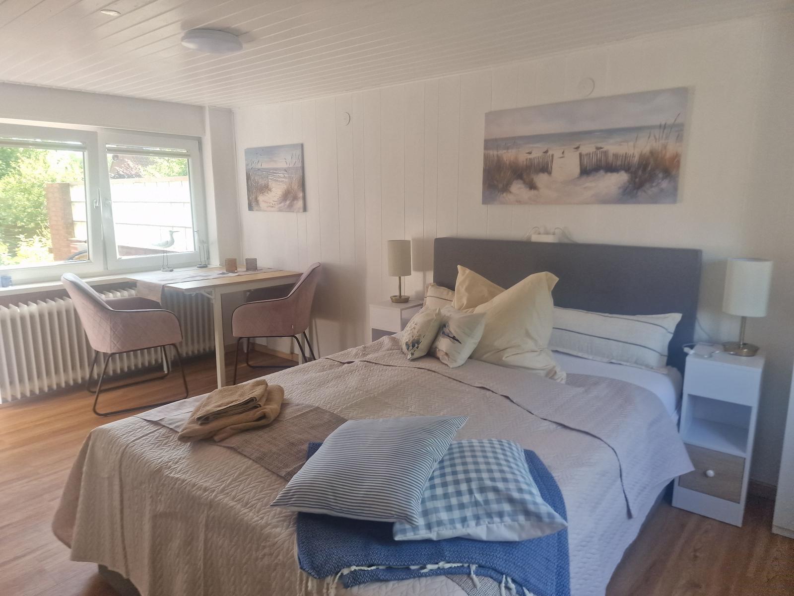 Schlafzimmer mit Bett, Schreibtisch und Fenster. Wandbilder zeigen Strand.