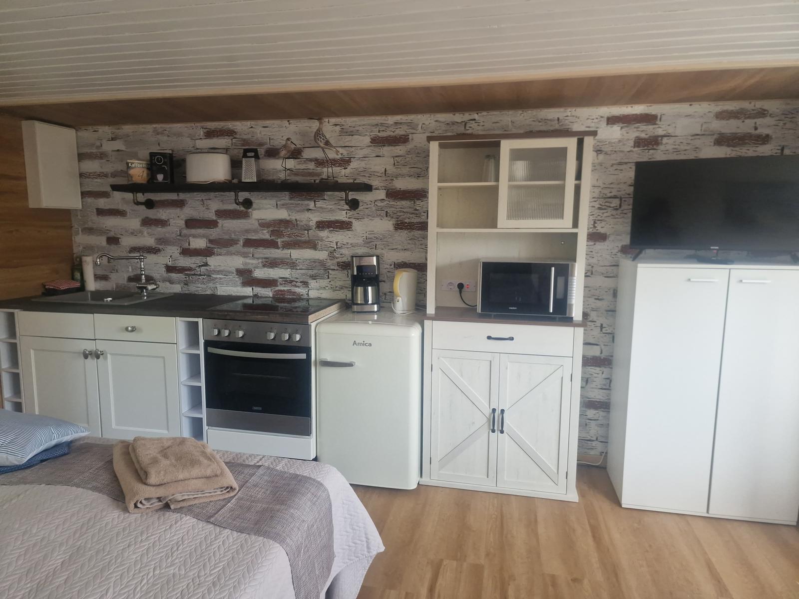 Küche mit Kühlschrank, Herd und Mikrowelle. Bett und Fernseher im Hintergrund.
