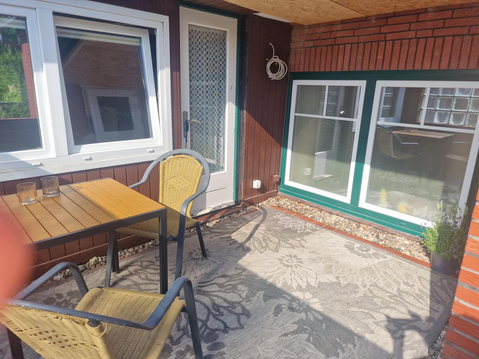 Terrasse mit Tisch, zwei Stühlen und Teppich. Hintergrund: Hauswand mit Fenstern und Tür.
