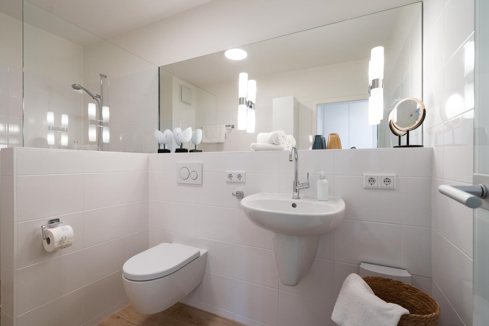 Badezimmer mit Toilette, Waschbecken und Dusche. Weiße Fliesen und Spiegel.