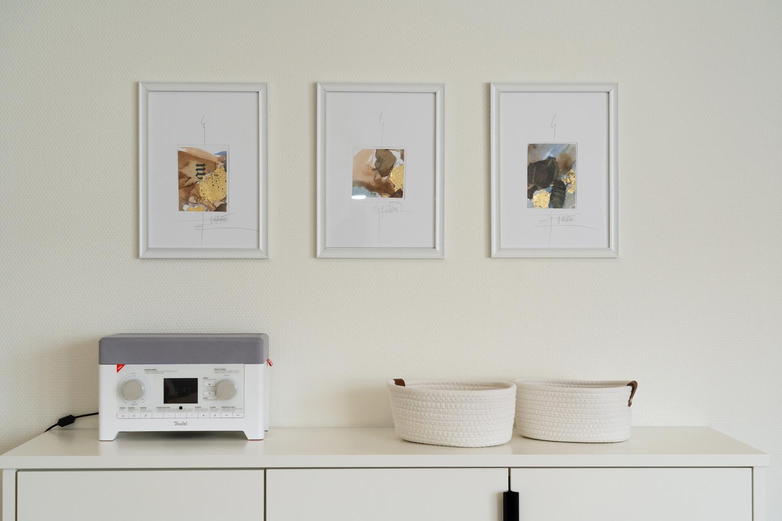 Drei gerahmte Kunstwerke hängen über einem weißen Schrank mit Korb und Audio-Gerät.