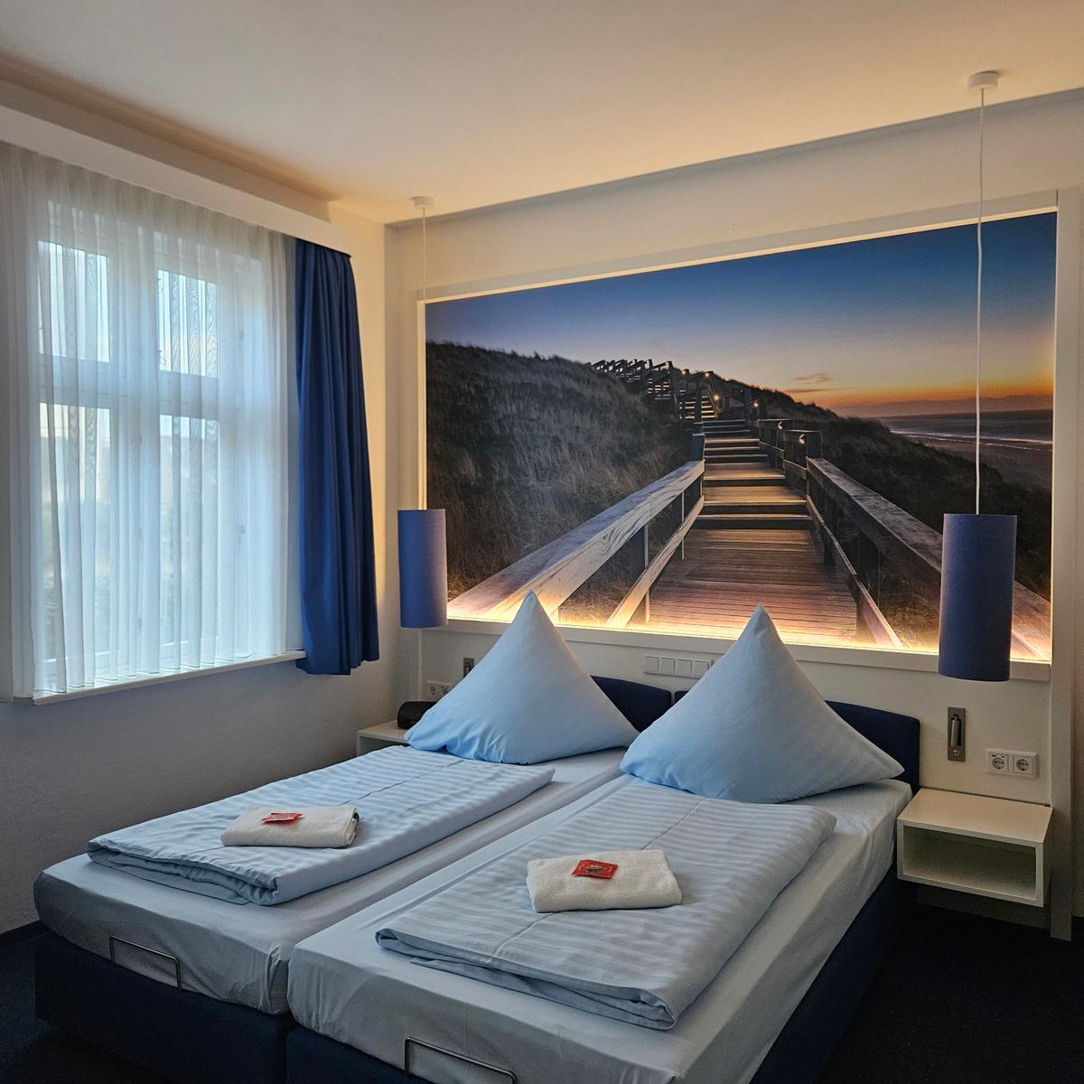 Zwei Betten mit blauen Kissen und Wandbild mit Strandweg.
