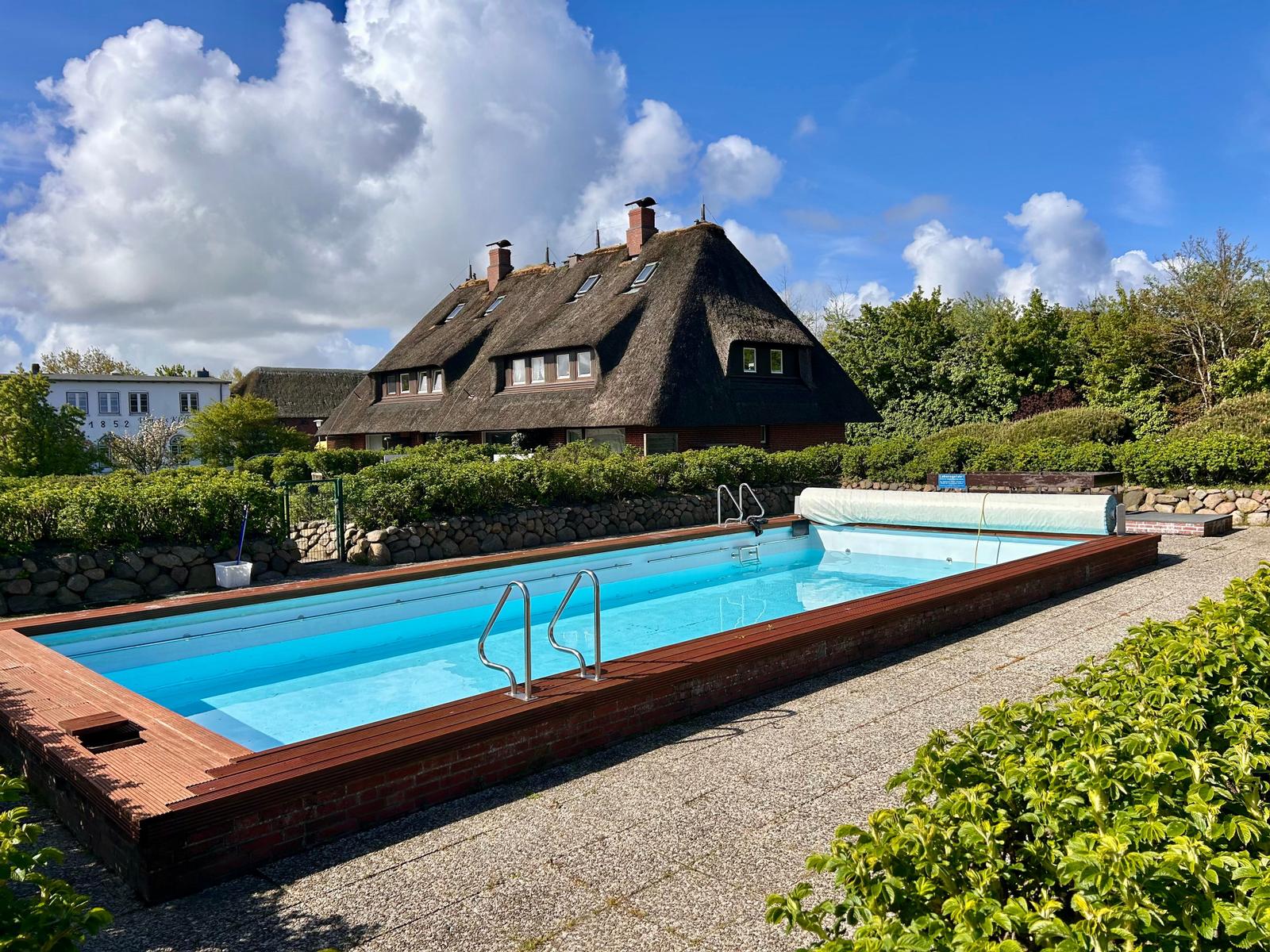 Pool mit Holzrand und Schwimmsteg vor einem Dachhaus mit Strohdach.