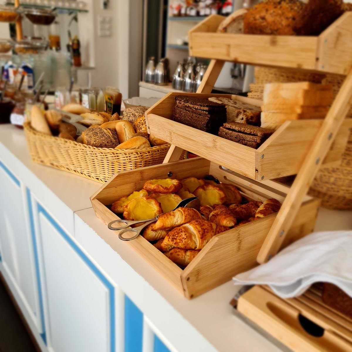 Frühstücksbuffet mit Brot und Croissants in Holzkisten auf einer weißen Theke.