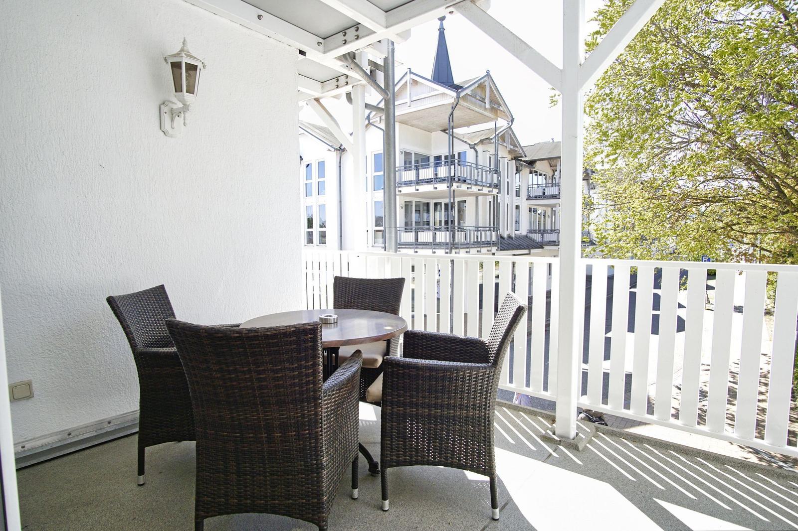 Terrasse mit Tisch und vier Stühlen, weißer Geländer, Blick auf Gebäude und Bäume.