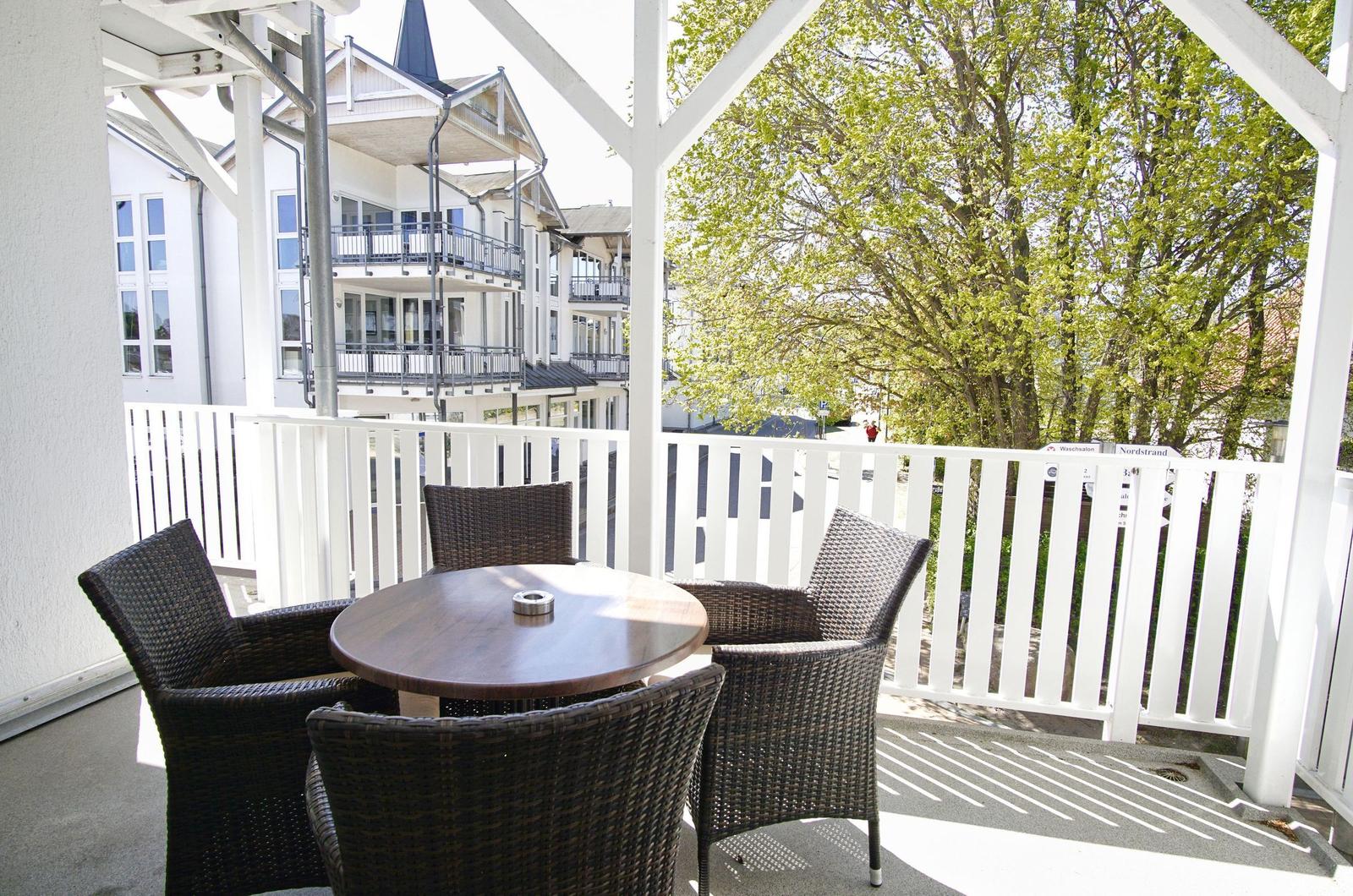 Terrasse mit runden Tisch und vier Stühlen, weißer Geländer und Blick auf Gebäude und Bäume.
