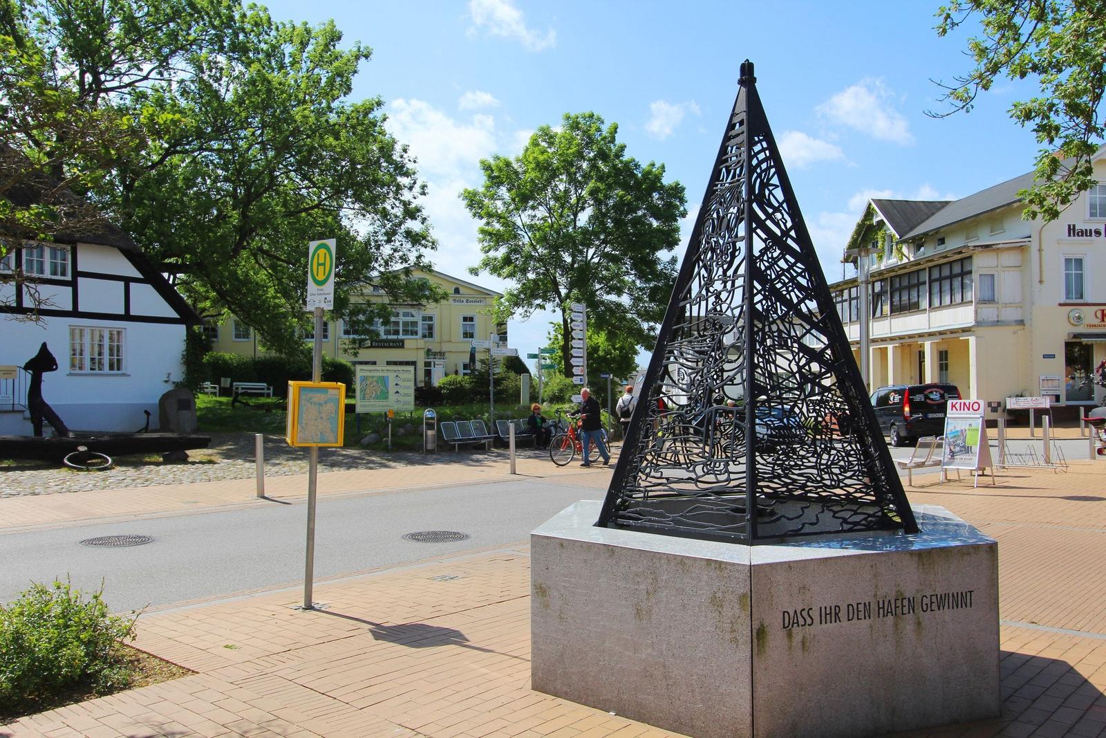 Drahtiger Turm auf Steinsockel mit Inschrift. Hintergrund: Straßenbild mit Geschäften und Bäumen.