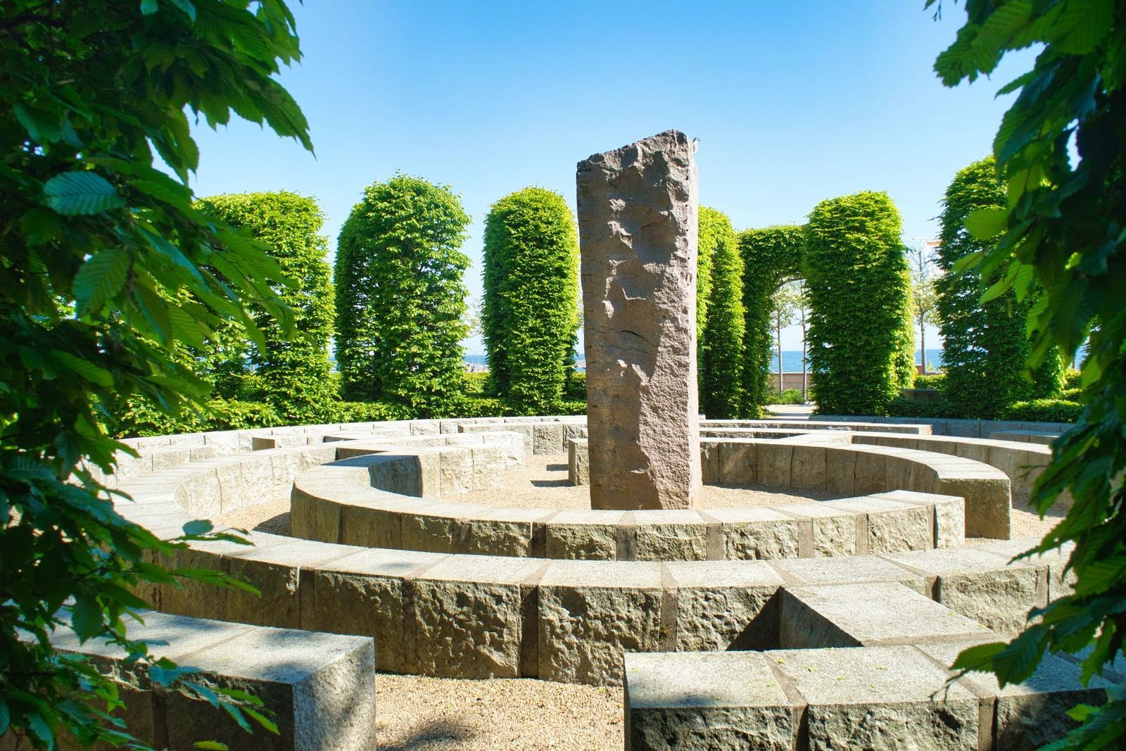 Steinmauer-Labyrinth mit Zentralpfeiler und grünen Bäumen im Hintergrund.